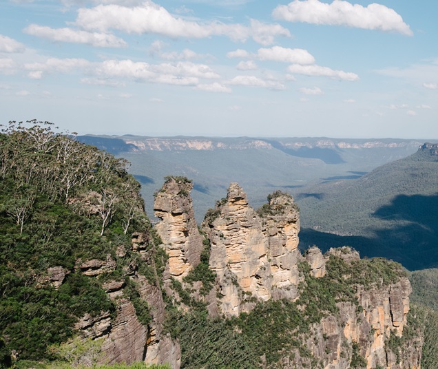 Three Sisters Blue Mountains Three sandstone rock formations rising from a forested cliff with distant blue mountains and a bright sky above.